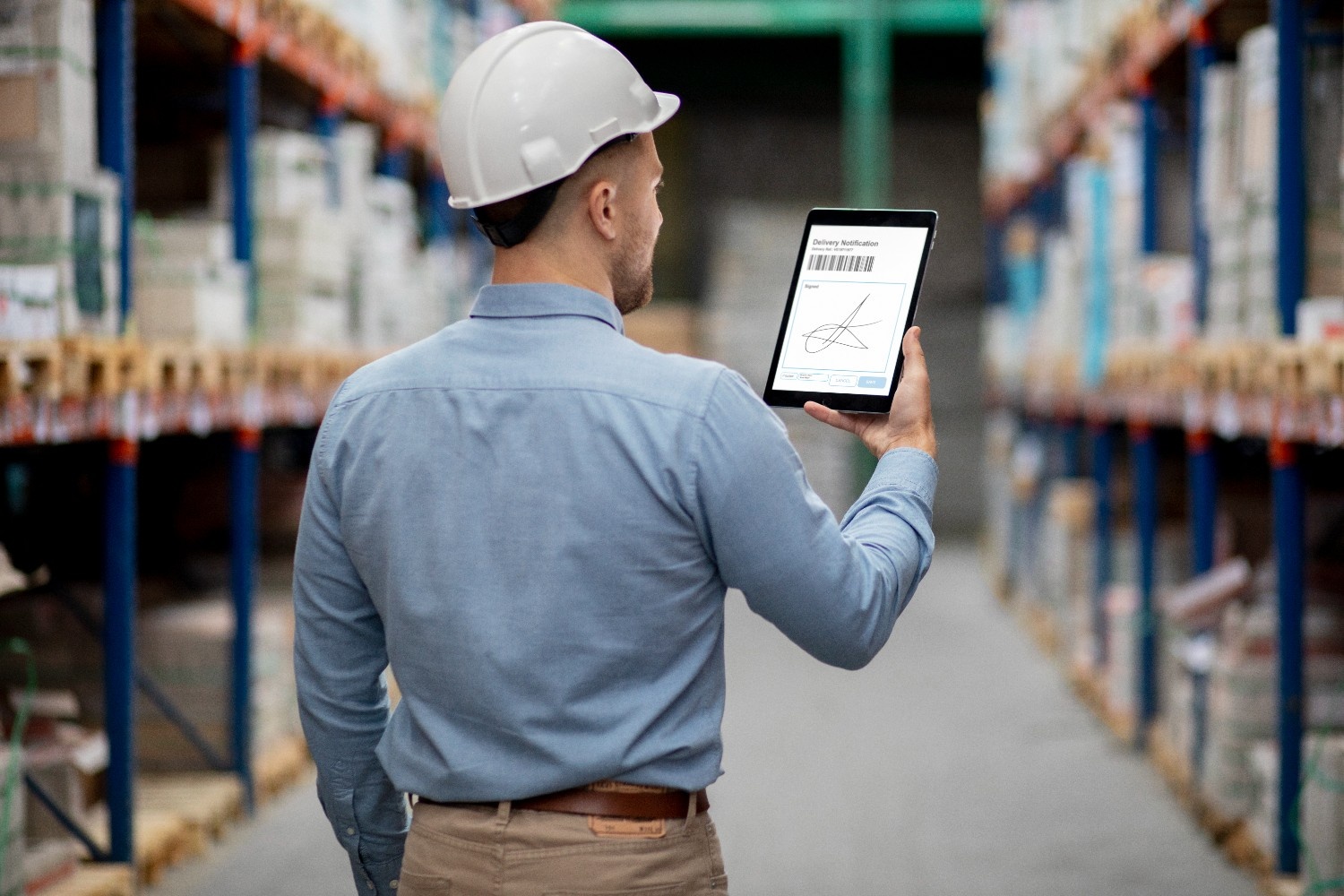 Warehouse worker wearing a white hard hat and blue shirt reviewing a delivery notification with barcode and digital signature on a tablet while standing in an industrial warehouse aisle
