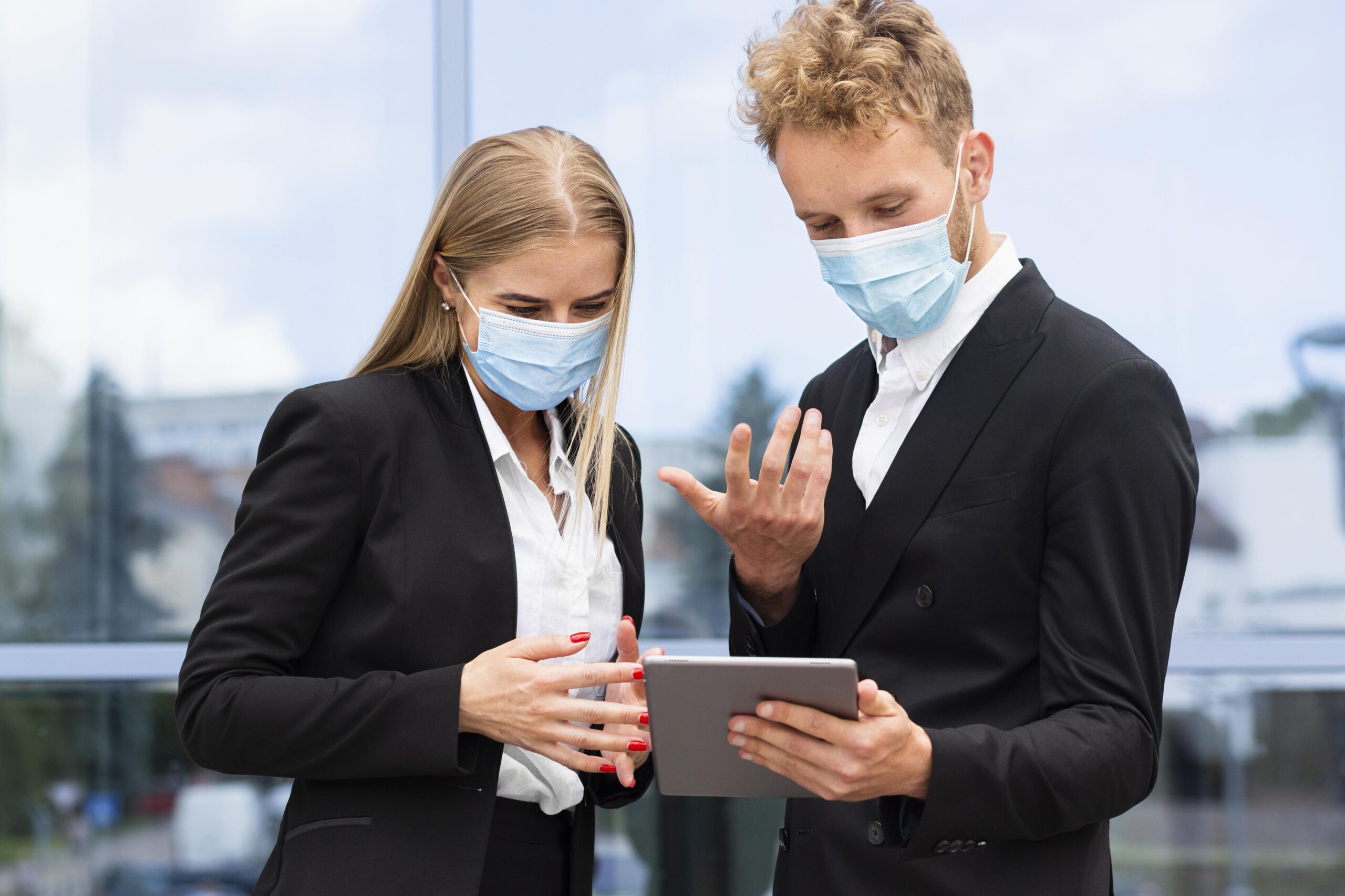 Two business professionals in black suits and blue face masks reviewing information on a tablet, representing e Signature in Insurance.