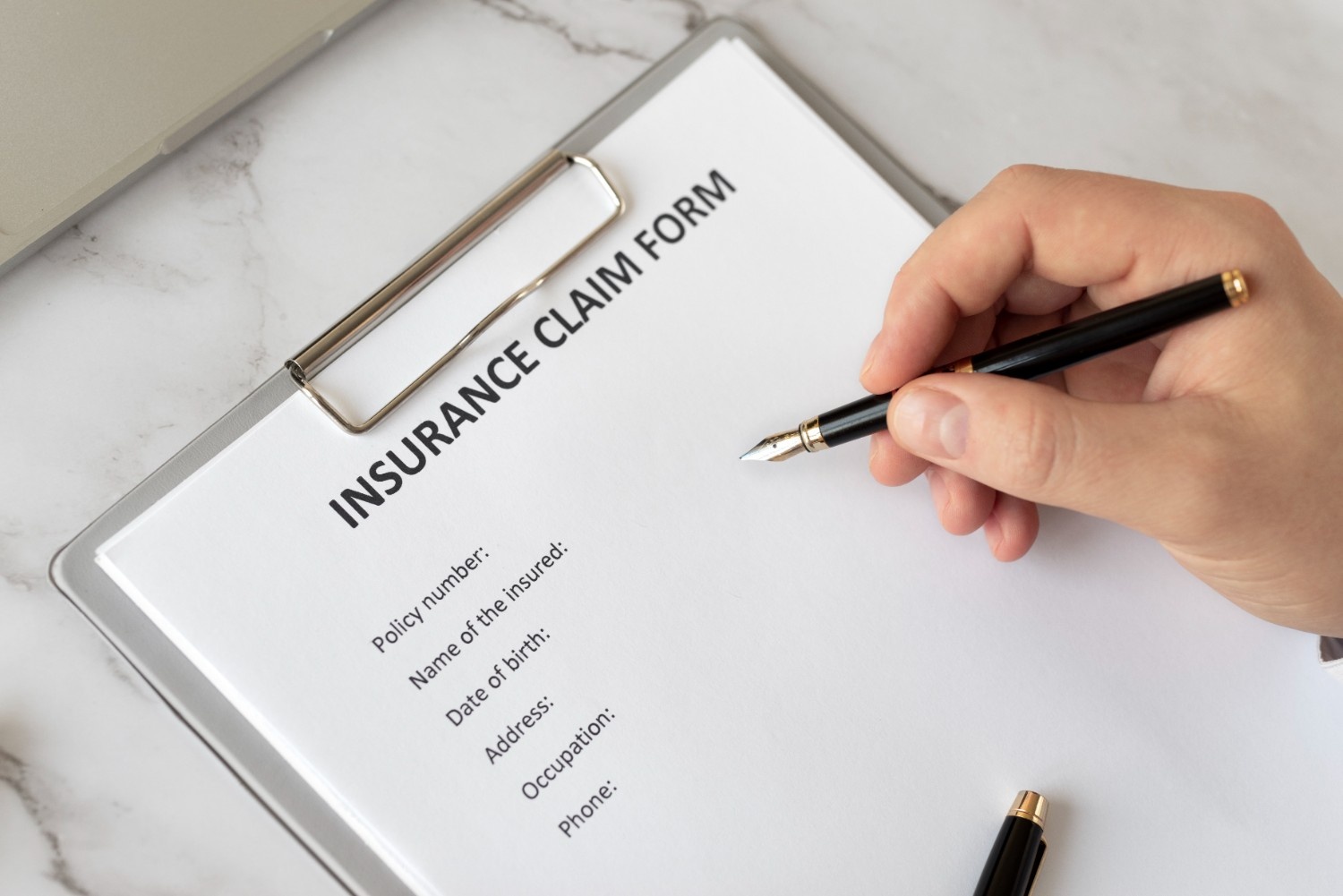 Hand holding fountain pen over insurance claim form on clipboard, with visible fields for policy number, name of insured, date of birth, address, occupation, and phone number on white paper with marble surface background