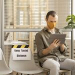 Man wearing orange face mask sits in modern office waiting area using tablet, with social distancing signs on adjacent chairs and monstera plant nearby
