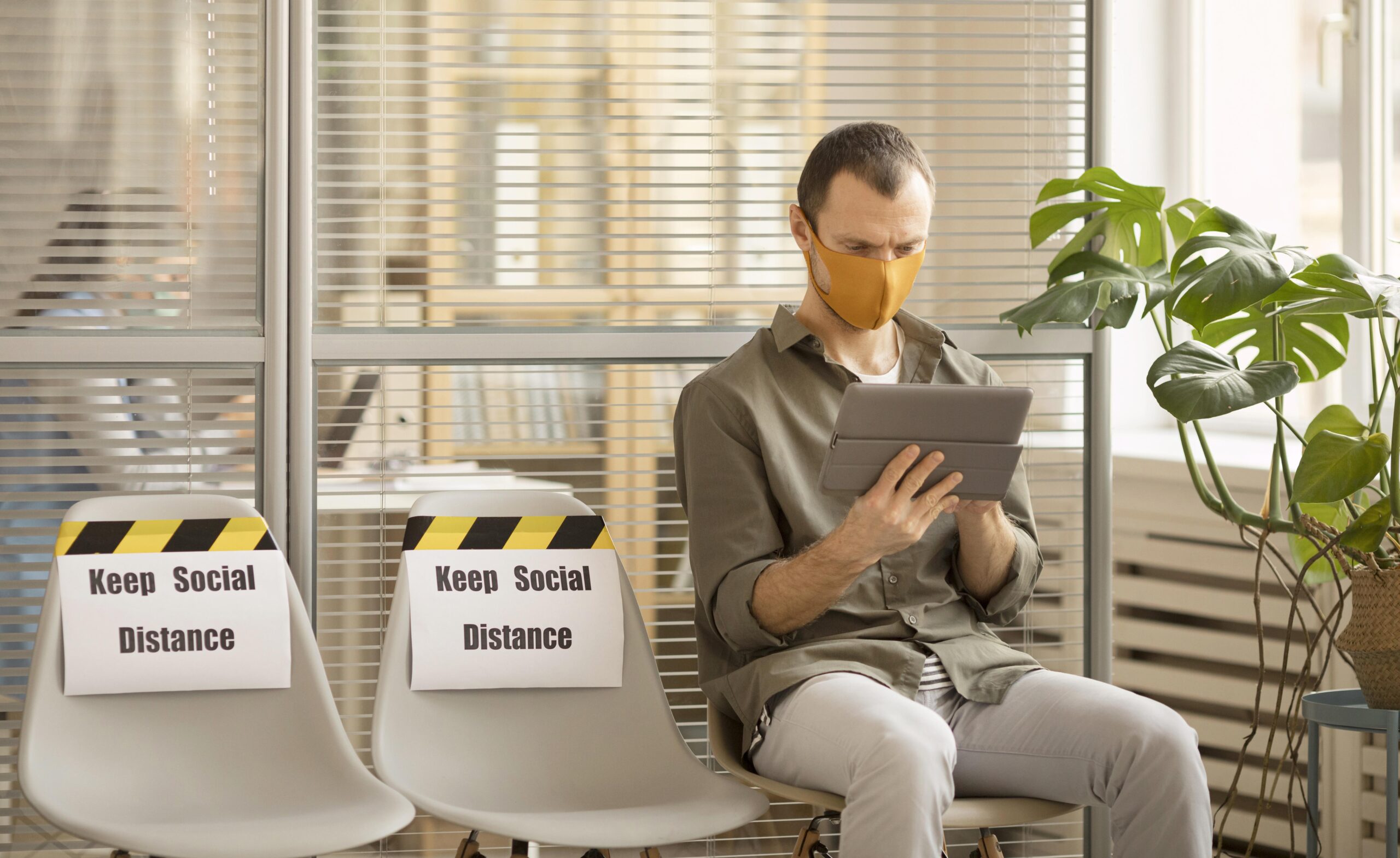 Man wearing orange face mask sits in modern office waiting area using tablet, with social distancing signs on adjacent chairs and monstera plant nearby