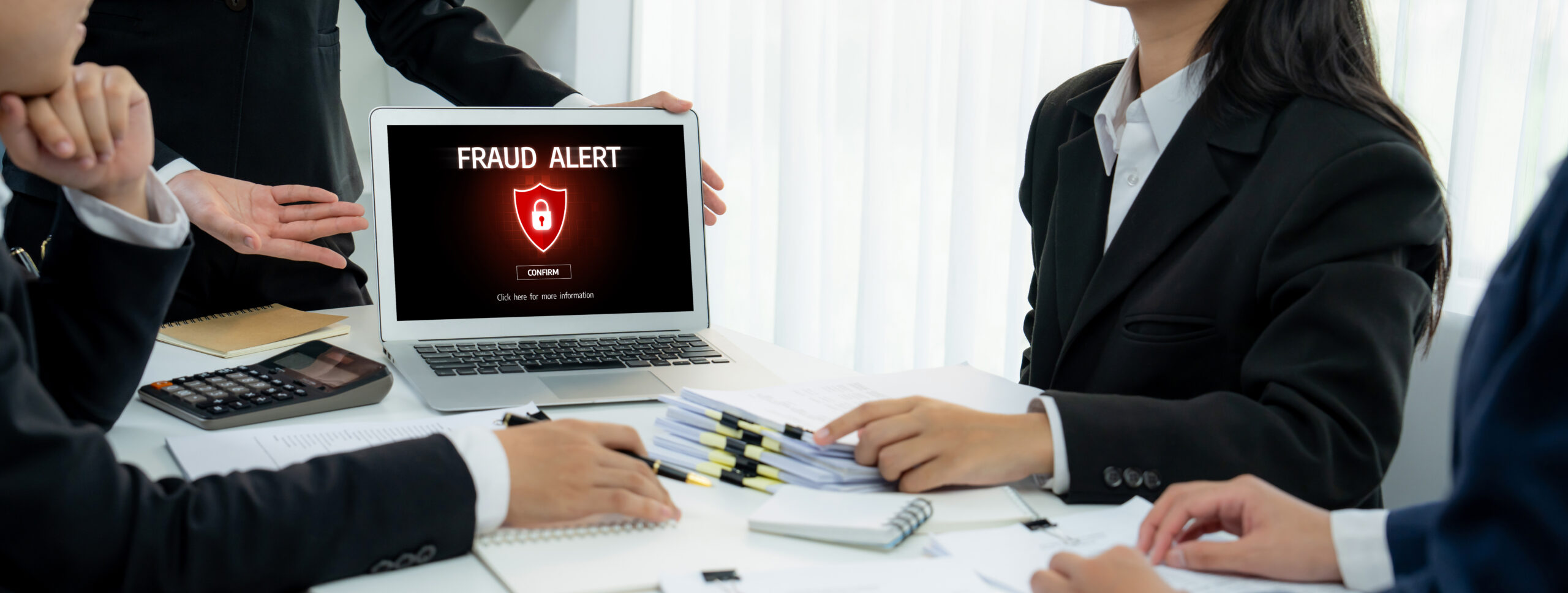 Business professionals in suits gathered around a laptop displaying a fraud alert warning with a red shield and lock icon on screen, reviewing documents and charts during a security meeting