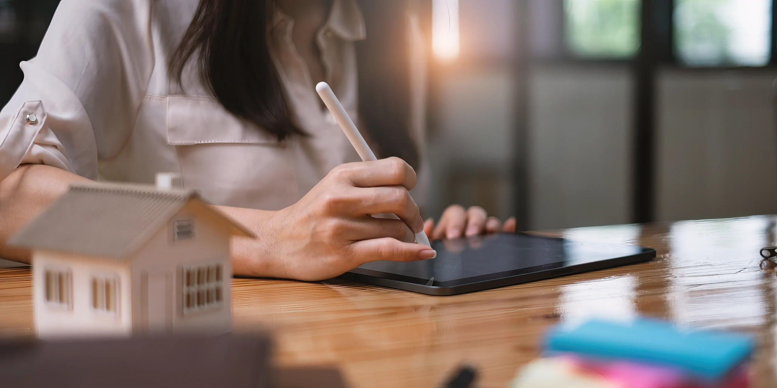 Real estate agent in tan blazer presenting a property contract on clipboard with pen, alongside miniature house model with red roof and calculator on white desk