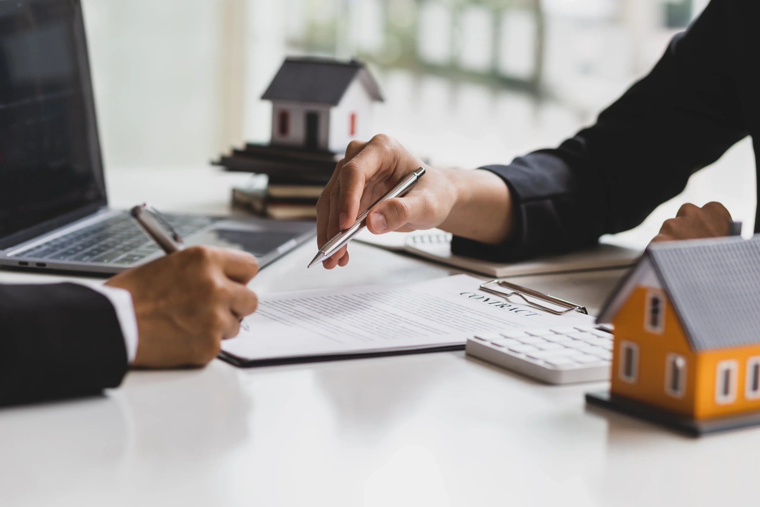 Real estate agent or mortgage broker consulting with client at desk, using tablet and calculator with house model and contract documents present, during property transaction discussion