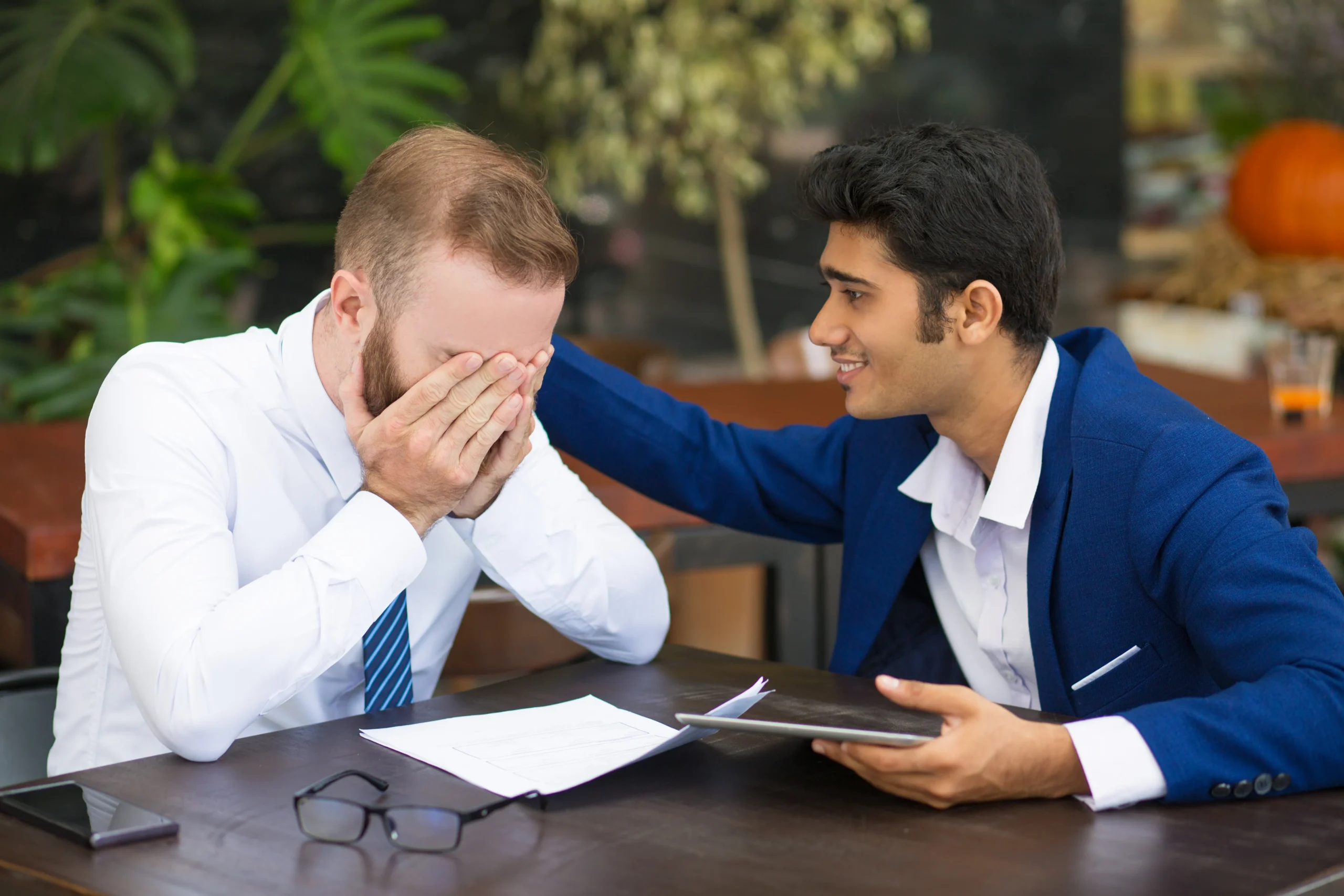 Business colleague in blue suit comforting distressed coworker in white shirt during difficult conversation at office table with documents and tablet.