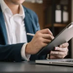 Business professional in dark suit using stylus pen on tablet in office environment with blurred colleagues in background.