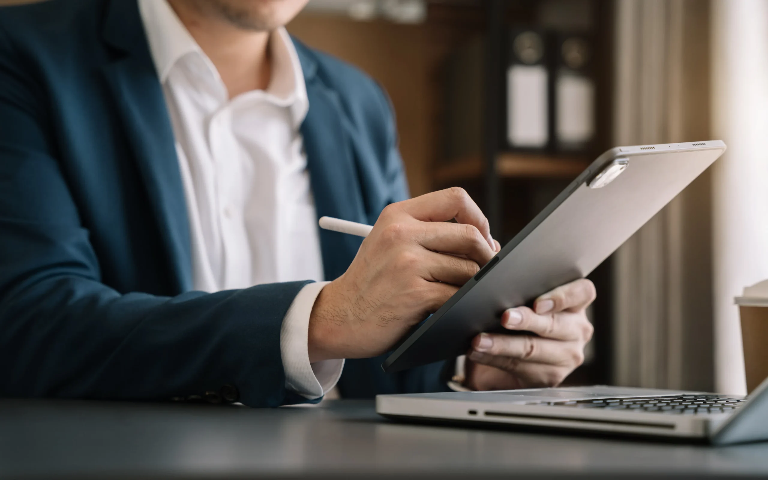 Business professional in dark suit using stylus pen on tablet in office environment with blurred colleagues in background.