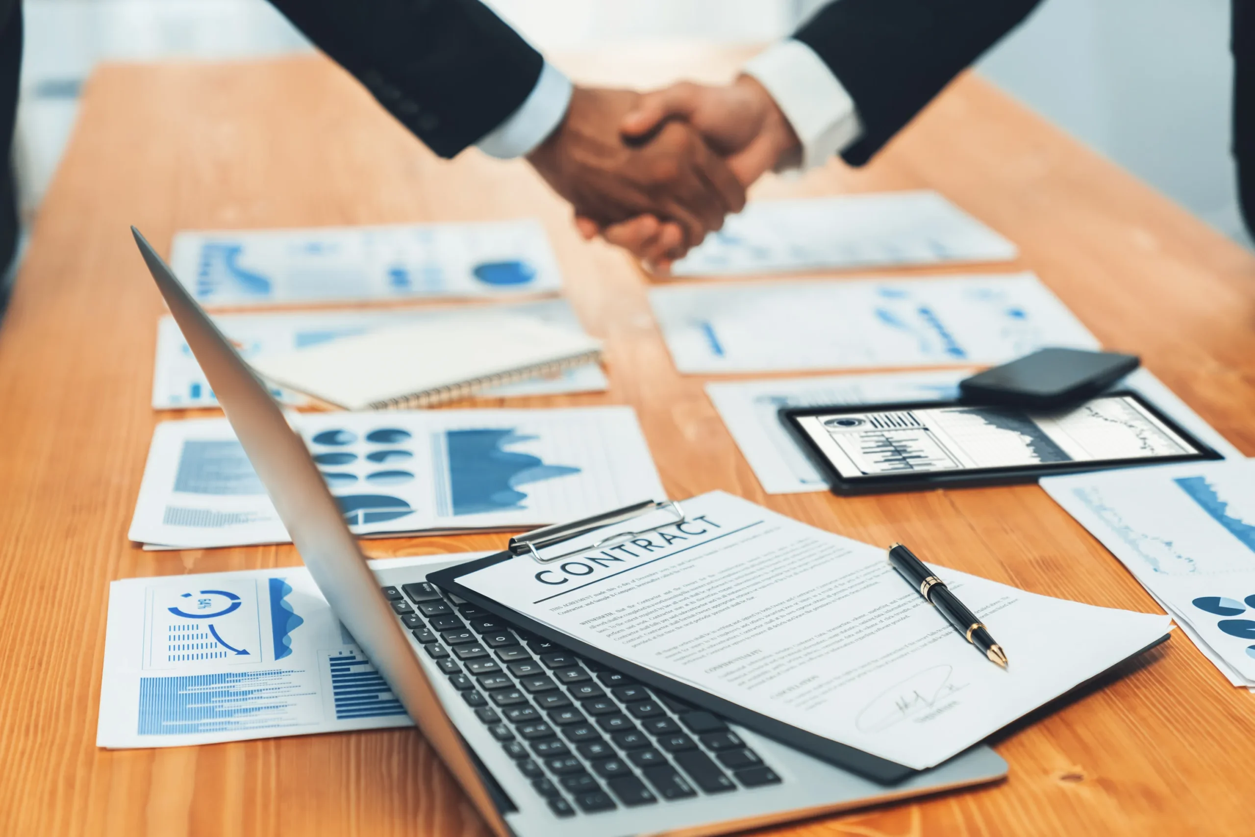 Two business professionals in suits shaking hands over desk covered with contract document, business charts, graphs, laptop, tablet, and pen, sealing a business deal