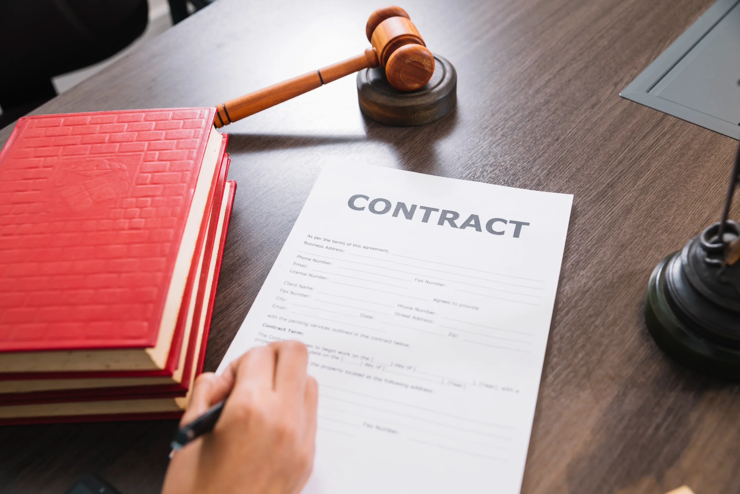 Person signing legal contract document on desk with wooden gavel, red law books, and laptop in background