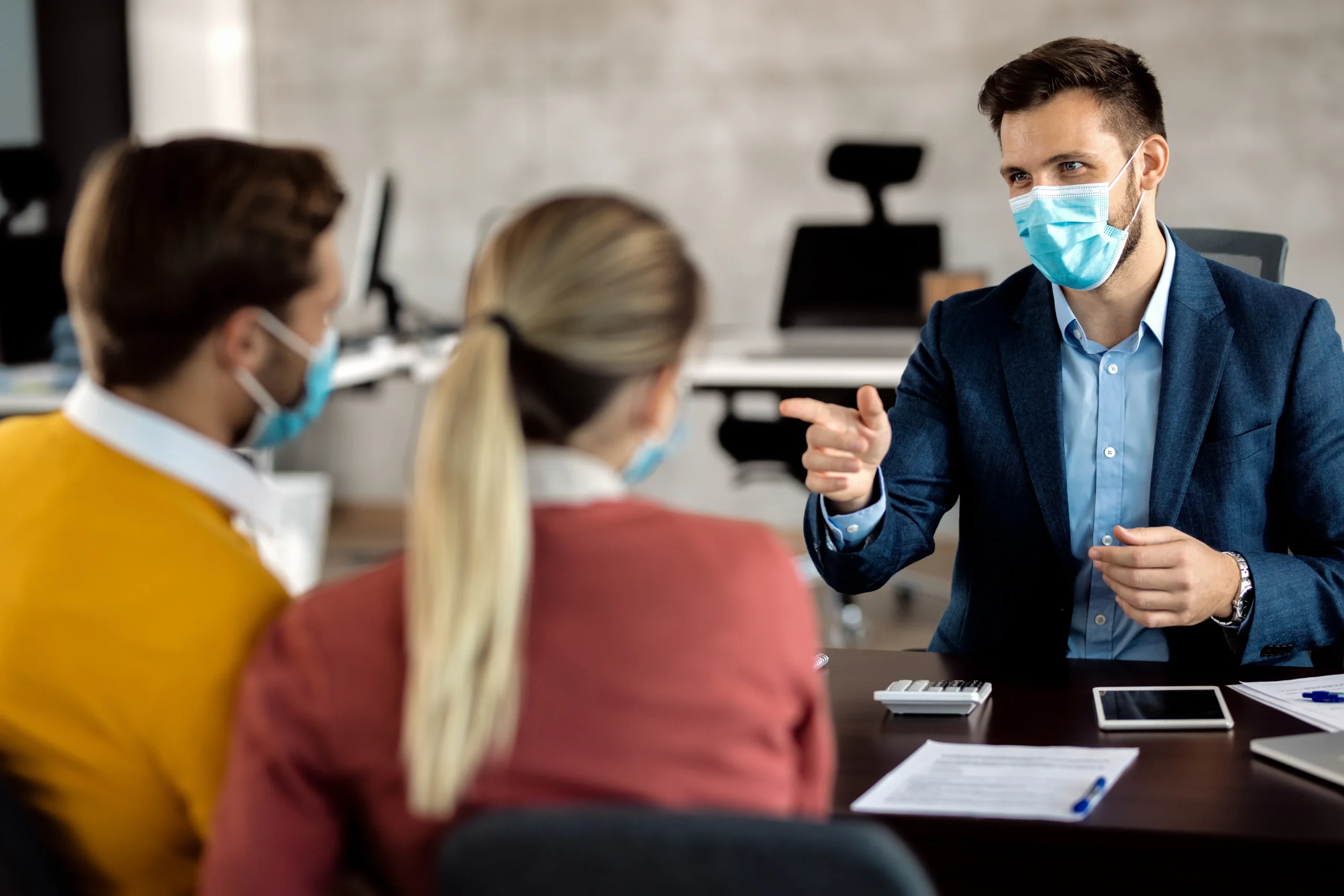 Business professional wearing face mask handing pen to client for document signing across desk during COVID-19 pandemic