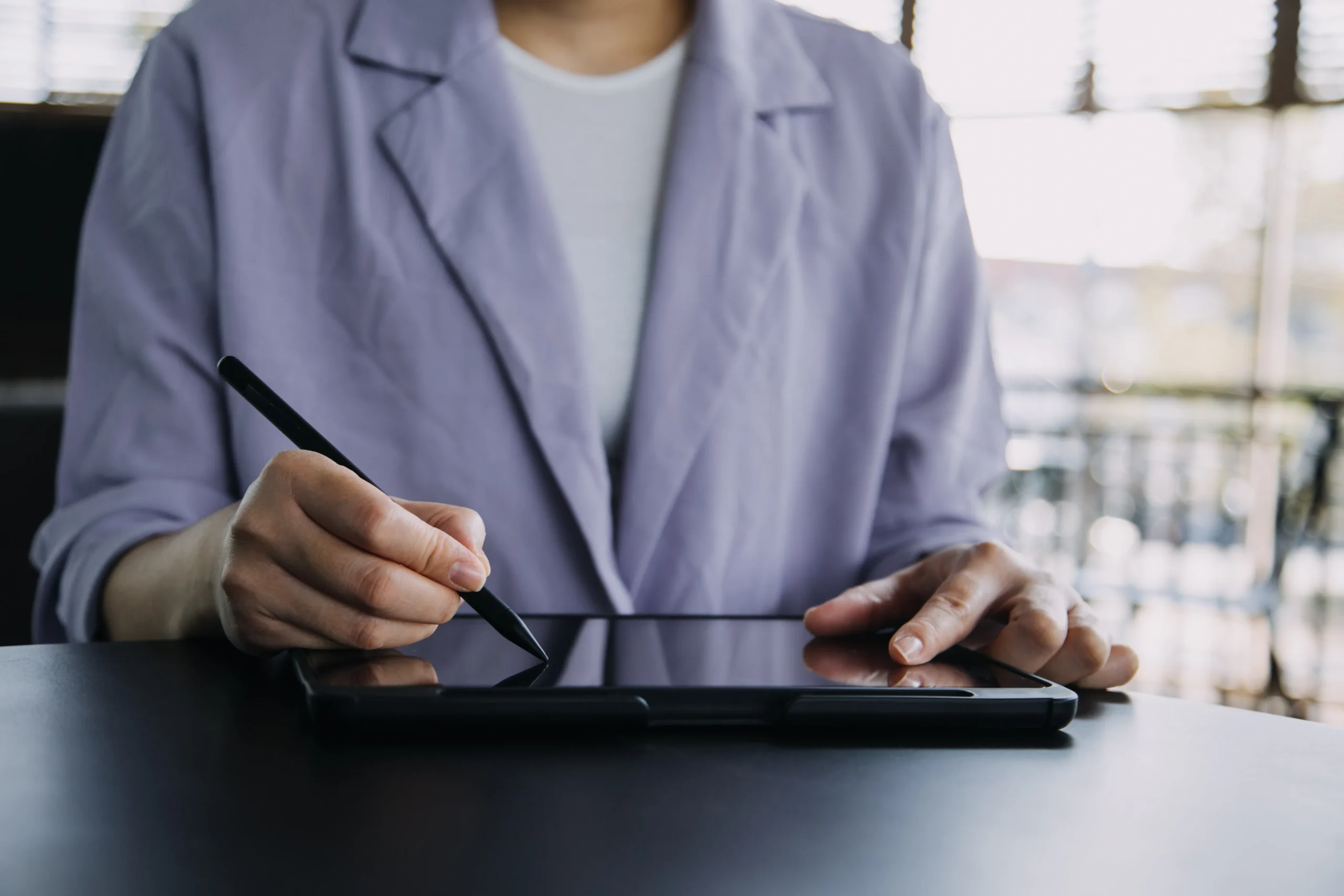 Professional in gray blazer using stylus pen on digital tablet at desk in bright office