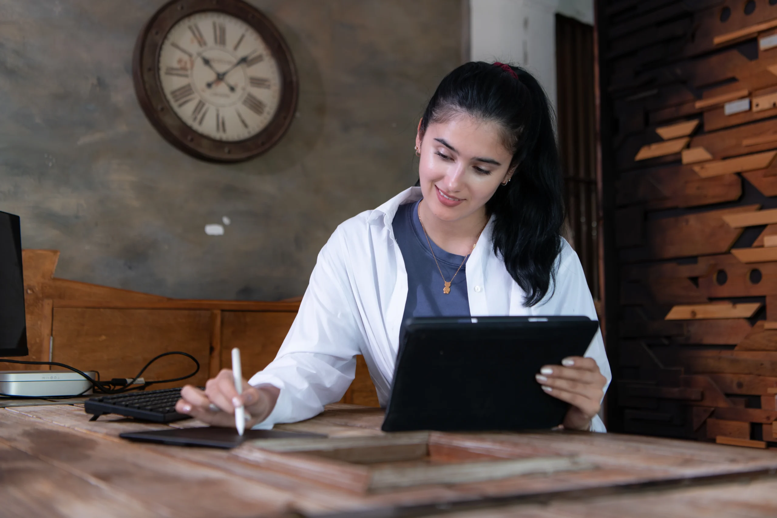 Woman in white cardigan and blue shirt working at a wooden desk, holding a tablet in one hand while writing notes with a stylus in the other, with a vintage wall clock and wooden shelving visible in the rustic background
