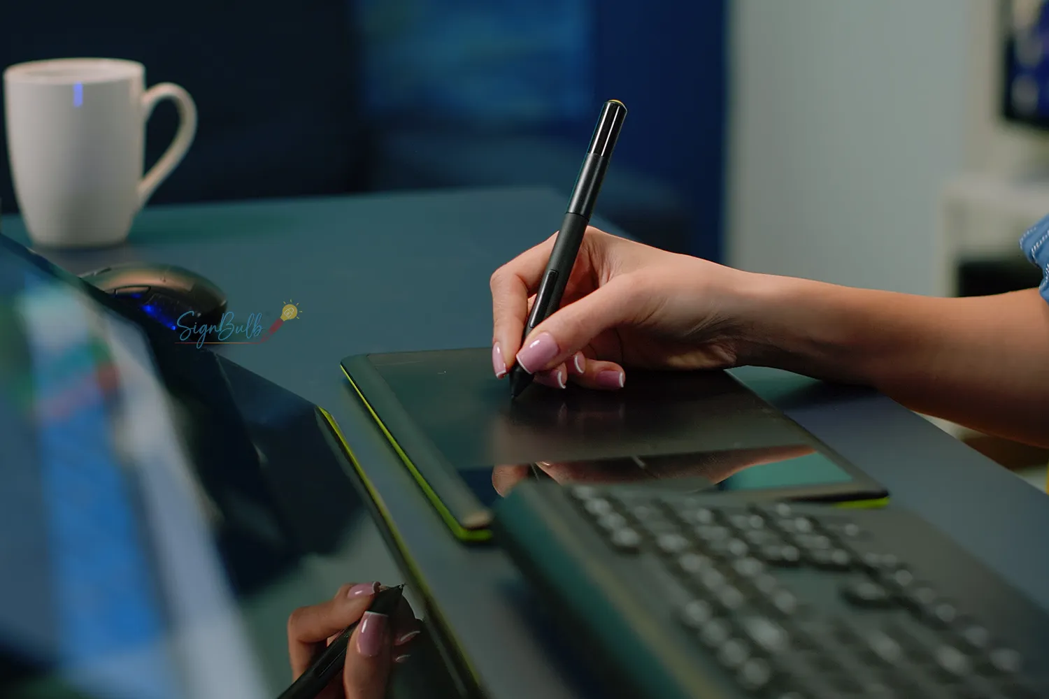 Person drawing on a graphics tablet with a stylus at a desk, laptop nearby and a white mug in the background, showing a creative workspace.