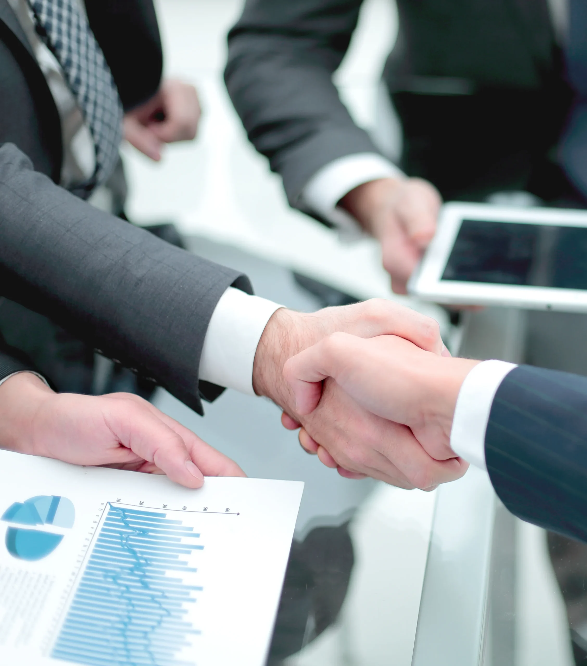 Business professionals shaking hands over a glass desk during a successful digital contract signing session, with one holding a tablet and another holding a data report.