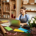 Woman in a flower shop talks on her phone at a cluttered craft desk with colorful papers and a laptop.