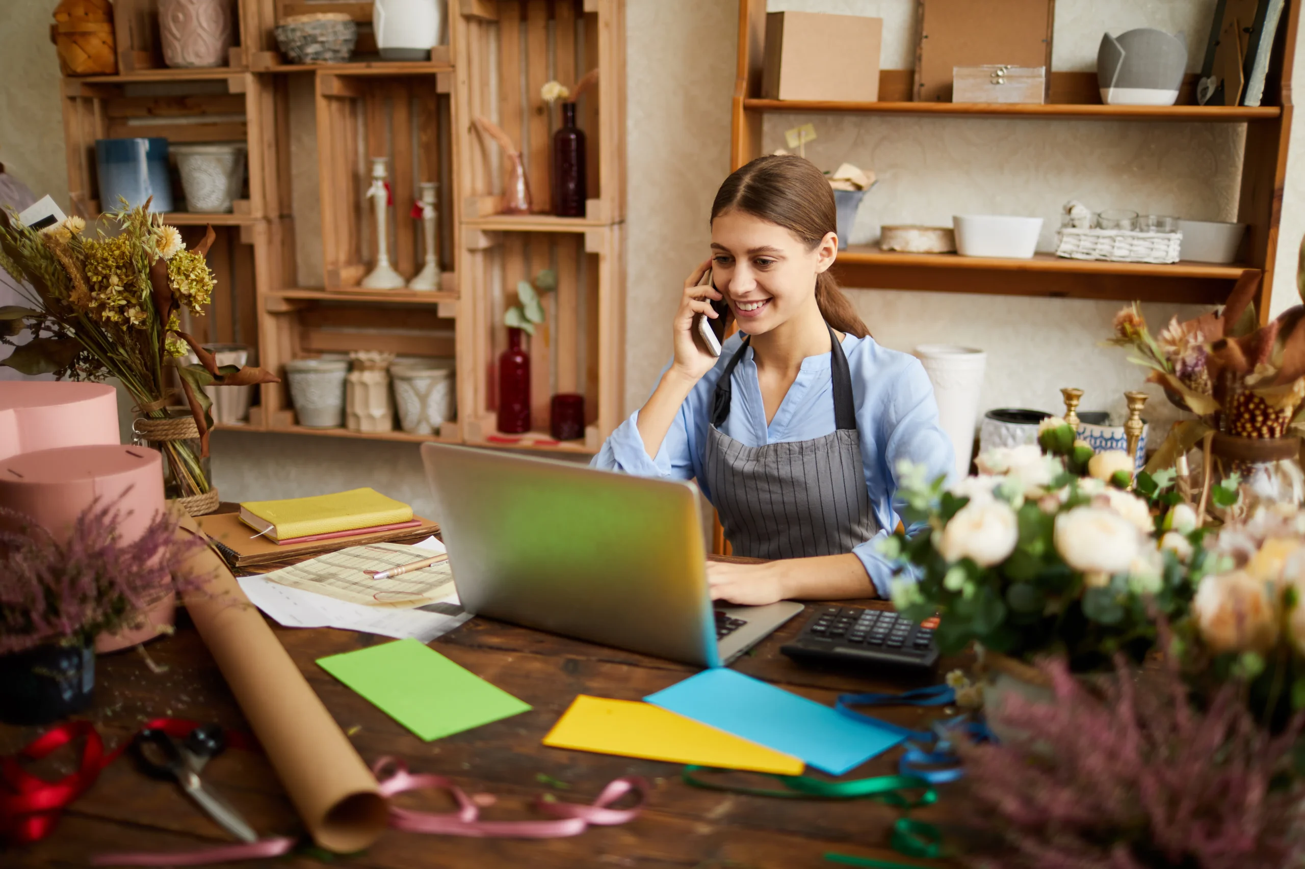 Woman in a flower shop talks on her phone at a cluttered craft desk with colorful papers and a laptop.